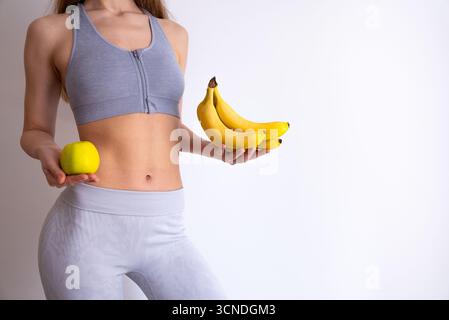 Junge Frau in grauer Aktivkleidung, die Banane und Apfel hält. Gesunde Ernährung und Fitness-Lifestyle-Konzept für Ernährung, Ernährungsplanung und Wellness. Stockfoto