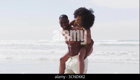 Mann, der eine Frau auf dem Rücken in flachem Meereswasser am Strand trägt, mit Badehosen, gelben Badeanzügen Stockfoto