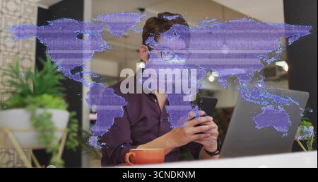 Mann trägt ein burgunderrotes Hemd, das Smartphone im Büro mit Laptop-Pflanzbecher und Karten-Overlay durchblättert Stockfoto