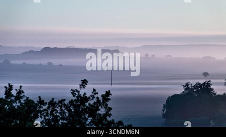 Sonnenaufgang über der französischen Landschaft mit sanft geschwungenen Feldern, verstreuten Bäumen und Morgennebel, gebadet in der Morgensonne Stockfoto
