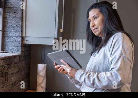Asiatische Frau, die Tablette von der Küchenfensterbank unter dem Schrank neben dem Papierhandtuchhalter benutzt Stockfoto