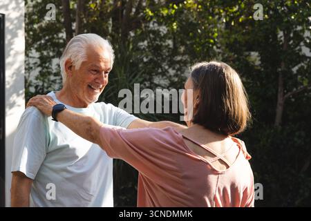 Seniorenpaar tanzt im sonnendurchfluteten Garten mit einem Mann in Smartwatch und einer Frau in rosa Bluse Stockfoto