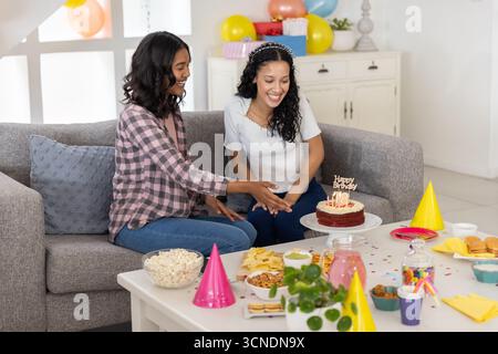 Vielfältige Mutter und Teenager-Tochter sitzen zu Hause auf der Couch und feiern Geburtstag mit Kuchen Stockfoto