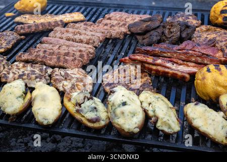 Auswahl an gegrilltem Fleisch und gerösteten Kartoffeln auf einem Grill. Das Bild zeigt die Texturen und Farben der Speisen, einschließlich gegrillter Würstchen und Schweinefleisch Stockfoto