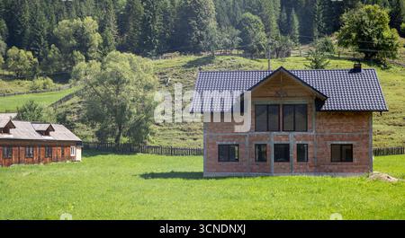 Ländliches Backsteinhaus und ein altes traditionelles Holzhaus befinden sich auf einem grünen Hügel mit üppiger Waldkulisse. Das Bild erfasst den Kontrast zwischen alten und Stockfoto