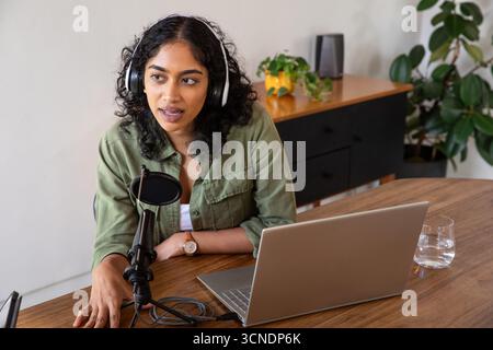 Indische Frau nimmt Podcast am Heimarbeitsplatz mit Mikrofon, Laptop und Kopfhörern auf Stockfoto