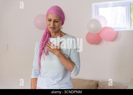 Ältere Frau, die zu Hause steht, trägt rosa Kopftuch und gestreiftes Oberteil, das die Brust berührt Stockfoto