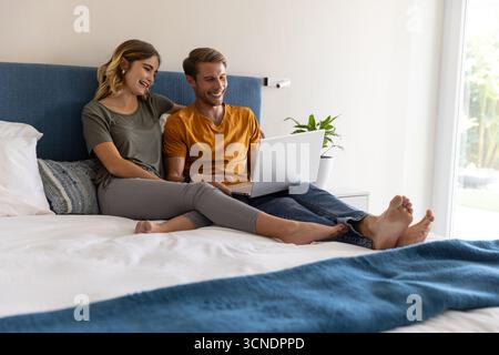 Ein Paar, das sich im Schlafzimmer gegen das blaue Kopfteil lehnt, einen Laptop teilt und sich auf Laken ausruht Stockfoto