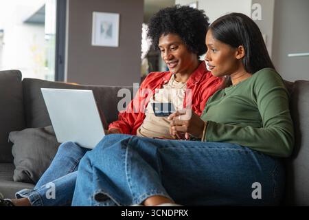 afroamerikaner und asiatisch-indisches Paar, das zu Hause auf der Couch mit Kreditkarte auf dem Laptop surft Stockfoto