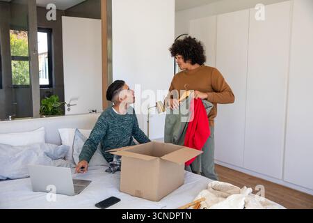 Männliche Mitbewohner packen Kleidung aus Pappkarton auf dem Bett und Sortierbügel im gut beleuchteten Schlafzimmer Stockfoto