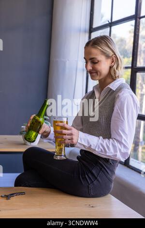 Frau, die am Tisch am Fenster sitzt und aus der Flasche in ein hohes Glas in der Nähe einer kleinen Schüssel strömt Stockfoto