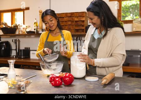 Asiatische Tochter und hispanische Seniormutter in Schürzen, die Mehl gesiebt und auf der Kücheninsel ein Glas hält Stockfoto