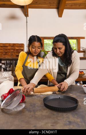 Vielfältige Mutter und Tochter in Schürzen rollender Teig mit Nudelnadel auf der Arbeitsplatte in der Küche Stockfoto