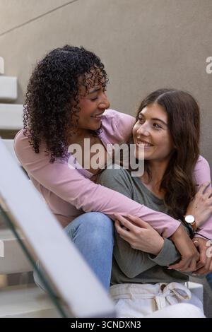 Verschiedene weibliche Paare sitzen auf Betontreppen, umarmen und lächeln mit Handlauf und Armbanduhr Stockfoto