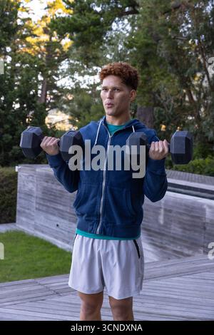 Mann mit Hoodie, der schwarze sechseckige Kurzhanteln auf Holzdeck an Wand hebt Stockfoto