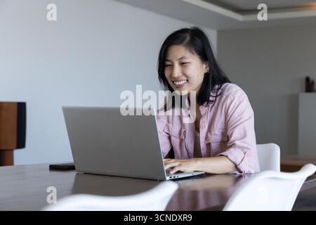 Asiatische koreanische Frau, die auf silbernem Laptop im Esszimmer mit Smartphone, Kopierraum tippt Stockfoto