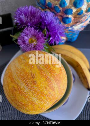 Eine gelbe, reife, duftende Melone und Bananen auf einer Platte mit violetten Astern, ein herbstliches Stillleben Stockfoto