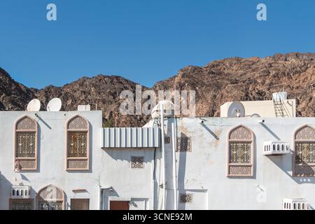 Oman - Muscat - Altstadt - sonnendurchflutete weiße Fassaden mit verzierten Fenstern unter gezackten braunen Hügeln. Stockfoto