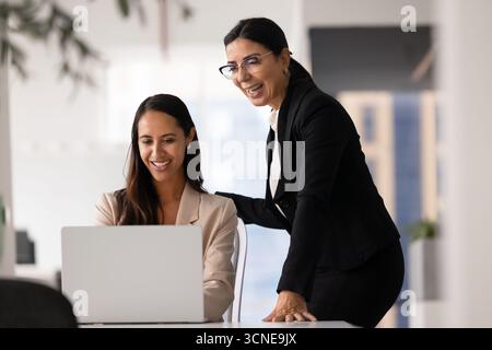 Glückliche, vielfältige weibliche Kollegen, die am Arbeitsplatz stehen und sitzen Stockfoto