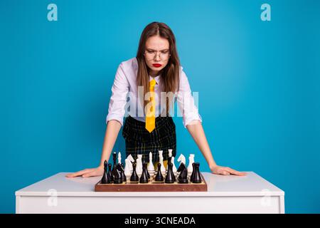 Junge Frau konzentriert sich auf Schachspielstrategie am Tisch mit blauem Hintergrund und zeigt eine professionelle und lehrreiche Umgebung Stockfoto