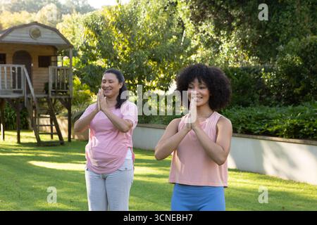 Hispanische Mutter und afroamerikanische Tochter üben Yoga im Garten mit Holzspielhaus Stockfoto
