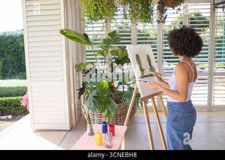afroamerikanerin, die botanische Formen an der Staffelei auf der überdachten Veranda mit Farbgläsern malte Stockfoto