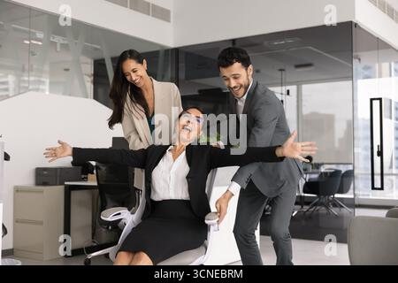 Positive Angestellte schieben Bürosessel mit lachender Chefin Stockfoto