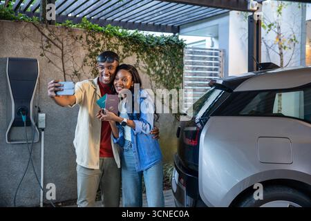 Afroamerikanisches Paar, das unter Pergola steht und Selfie macht, mit Pässen neben dem EV-Ladegerät Stockfoto