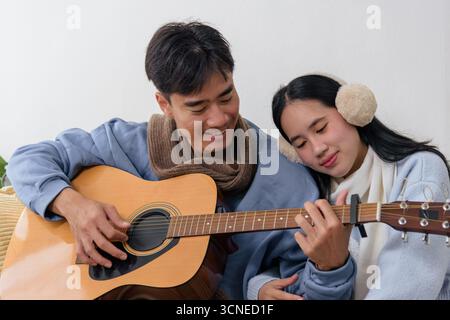 Wintertag drinnen, fröhliches junges Paar genießt gemütliches Heim mit Musik, Mann spielt Gitarre, Frau Stockfoto