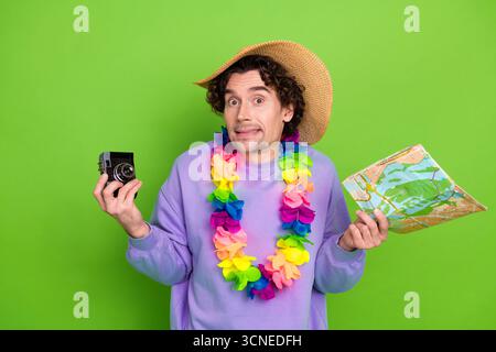Junger Mann mit Kamera und Karte, mit tropischer lei-Halskette und Strohhut, neugierig vor grünem Hintergrund Stockfoto