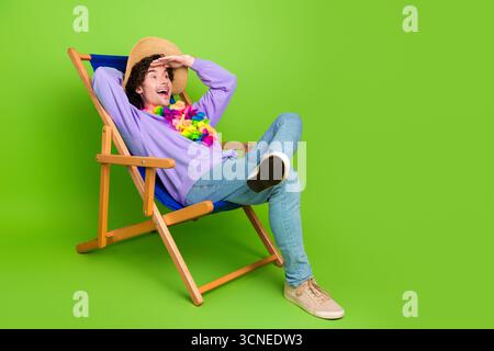 Schöner Mann, der sich auf einem Stuhl vor einem leuchtend grünen Hintergrund entspannt, in lässiger Kleidung und einem Strohhut trägt. Stockfoto