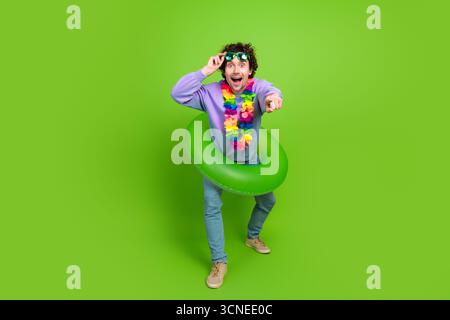 Fröhlicher junger Mann mit lockigen Haaren, der mit grünem Schwimmer auf lebendigem Hintergrund posiert und Aufregung ausdrückt Stockfoto
