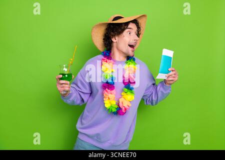 Aufgeregter junger Mann in trendiger Freizeitkleidung mit Ticket und Getränk vor einer lebendigen grünen Kulisse im Studio Stockfoto