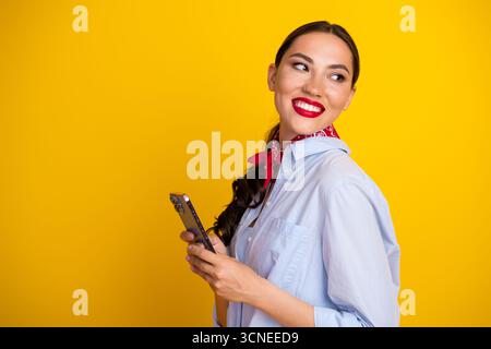 Lächelnde Frau, die ein Smartphone vor einem leuchtend gelben Hintergrund hält und Mode und lässige Eleganz zeigt Stockfoto