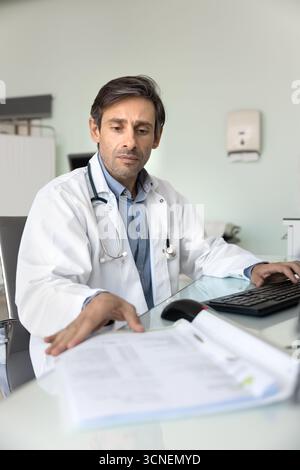 Männlicher Arzt, der mit Papierdokumenten am Schreibtisch am pc arbeitet Stockfoto