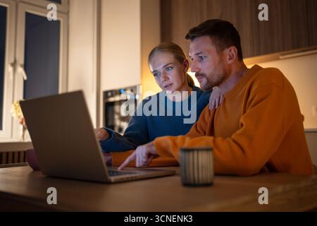 Engagierte Ehemann und Ehefrau, die nachts in der Küche im Internet einkaufen und soziale Medien nutzen Stockfoto