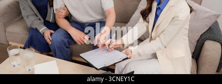 Ein professioneller asiatischer Immobilienmakler erklärt dem erwachsenen Paar die Vereinbarung. Kunden unterzeichnen einen Vertrag im Wohnzimmer zu Hause für sichere zukünftige Investitionen. Stockfoto