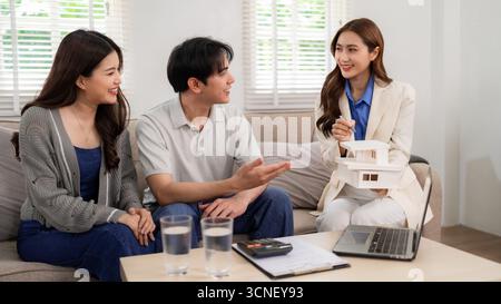 Freundliche asiatische Immobilienmaklerin erklärt dem jungen Paar in ihrer Wohnung das Hausmodell. Sie diskutieren mit Immobilienkaufinvestitionen und Hauseigentum Stockfoto