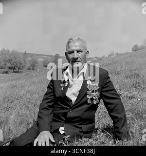 Ein Porträt eines Veteranen aus dem Zweiten Weltkrieg mit zahlreichen Orden und Medaillen auf der Brust, der am 9. Mai 1985 in Swiatohirsk, UdSSR, auf einem Feld vor natürlicher Kulisse saß. Dieses archivierte Schwarzweiß-Foto, das sowohl in seriöser als auch in lächelnder Version vorhanden ist, vermittelt tief die persönlichen Emotionen und Erinnerungen des Helden am Siegestag und zeigt sowohl die Trauer um die Vergangenheit als auch die Freude am friedlichen Leben Stockfoto