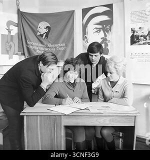 Junge Arbeiter nehmen in den 1970er Jahren an einem Treffen der Komsomol (Junge Kommunistische Liga) in der „Roten Ecke“ im AIZ-Werk in Sloviansk, UdSSR Teil Dieses Schwarzweiß-Archivfoto zeigt die Szene der Unterzeichnung eines wichtigen Dokuments vor dem Hintergrund sowjetischer Plakate und des Komsomol-Banners, das die Atmosphäre des ideologischen und sozialen Lebens der sowjetischen Jugend vermittelt Stockfoto