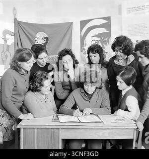 Junge Arbeiter nehmen in den 1970er Jahren an einem Treffen der Komsomol (Junge Kommunistische Liga) in der „Roten Ecke“ im AIZ-Werk in Sloviansk, UdSSR Teil Dieses Schwarzweiß-Archivfoto zeigt die Szene der Unterzeichnung eines wichtigen Dokuments vor dem Hintergrund sowjetischer Plakate und des Komsomol-Banners, das die Atmosphäre des ideologischen und sozialen Lebens der sowjetischen Jugend vermittelt Stockfoto