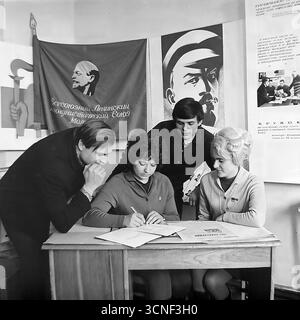 Junge Arbeiter nehmen in den 1970er Jahren an einem Treffen der Komsomol (Junge Kommunistische Liga) in der „Roten Ecke“ im AIZ-Werk in Sloviansk, UdSSR Teil Dieses Schwarzweiß-Archivfoto zeigt die Szene der Unterzeichnung eines wichtigen Dokuments vor dem Hintergrund sowjetischer Plakate und des Komsomol-Banners, das die Atmosphäre des ideologischen und sozialen Lebens der sowjetischen Jugend vermittelt Stockfoto