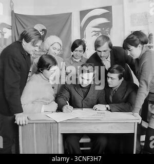 Junge Arbeiter nehmen in den 1970er Jahren an einem Treffen der Komsomol (Junge Kommunistische Liga) in der „Roten Ecke“ im AIZ-Werk in Sloviansk, UdSSR Teil Dieses Schwarzweiß-Archivfoto zeigt die Szene der Unterzeichnung eines wichtigen Dokuments vor dem Hintergrund sowjetischer Plakate und des Komsomol-Banners, das die Atmosphäre des ideologischen und sozialen Lebens der sowjetischen Jugend vermittelt Stockfoto