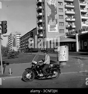 Ein sowjetischer Milizmann, ein Offizier der GAI (Staatliches Automobilinspektorat), sitzt während seines Dienstes an einer Straßenkreuzung in Sloviansk, ukrainische SSR, während der Feierlichkeiten zum Stadttag am 6. September 1985 auf seinem Patrouillenmotorrad. Im authentischen urbanen Hintergrund sind typische Wohnhäuser aus der Sowjetzeit mit riesigen Propagandamalereien dekoriert, darunter eines mit dem Slogan „Wir genehmigen die Politik der KPdSU!“, was eine kraftvolle und atmosphärische Momentaufnahme der Strafverfolgung und Ideologie in der späten UdSSR schafft Stockfoto