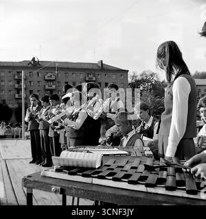 Ein Kindermusikensemble mit Melodika und Xylophon tritt auf einer Freiluftbühne in einem Wohnviertel während des Stadttags in Sloviansk, UdSSR, am 6. September 1985 auf. Dieses Vintage-Schwarzweiß-Foto fängt die authentische Atmosphäre eines lokalen sowjetischen Urlaubs und den Charme der Kreativität der Kinder ein. Die Vorstellung spielt vor dem Hintergrund typischer Apartmenthäuser und zeigt Feste auf Gemeindeebene Stockfoto