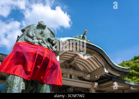 Vor dem reich verzierten Dach des Hokoku-Schreins in Osaka, Japan, steht eine Nahaufnahme einer Wächterlöwen-Statue mit rotem Tuch Stockfoto
