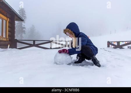 Schneemänner für Kinder an schneereichen Tagen. Ein Kind im Schnee, das einen Schneeball in der Nähe einer Holzhütte an einem nebeligen Tag rollt, mit verschwommenen Figuren im Stockfoto