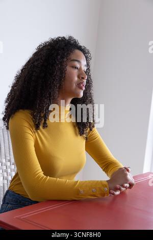afroamerikanerin mit Senfoberteil, die auf einem Korbstuhl am Tisch in der Zimmerecke sitzt Stockfoto