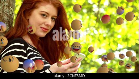 Rothaarige Frau, die sich gegen den Kofferraum lehnt und ein Smartphone hält, die Kamera im Park anstarrt, mit Emoji-Kugeln Stockfoto