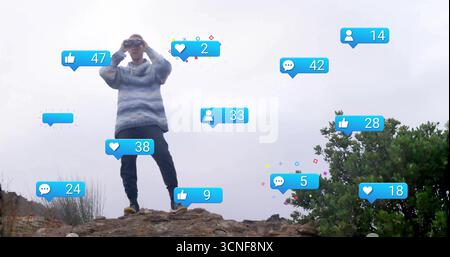 Scannen Sie den Wanderer in gestreiftem Hoodie mit Fernglas auf felsigem Felsvorsprung, mit schwebenden sozialen Symbolen Stockfoto